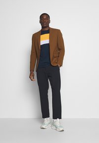 Brown blazer over a blue t-shirt with yellow and white stripes, paired with dark pants and light sneakers, standing against a plain background.