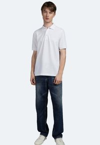 Polo bianco a maniche corte con bottoni frontali, abbinato a jeans in denim blu scuro e sneakers bianche. Design semplice e informale.