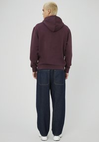 Jeune personne avec des cheveux blonds courts portant un sweat à capuche bordeaux, un jean bleu foncé à jambes larges et des baskets blanches, debout de dos.