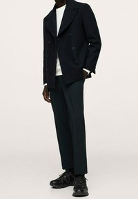 Granatowy wełniany płaszcz typu peacoat na dwa guziki, biały t-shirt z okrągłym dekoltem, dopasowane ciemne spodnie oraz czarne buty sportowe. Czysta, zorganizowana sylwetka.