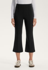 Pantalons noirs évasés en tissu extensible avec des coutures verticales le long de l'avant, associés à des baskets de couleur claire.