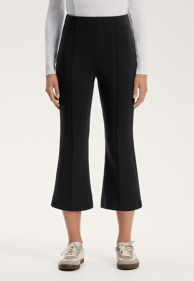 Pantalons noirs évasés en tissu extensible avec des coutures verticales le long de l'avant, associés à des baskets de couleur claire.