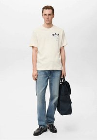 T-shirt a maniche corte color crema con testo blu "M.V.P.", abbinato a jeans azzurri chiari e scarpe di pelle nere, mentre tiene una borsa nera.
