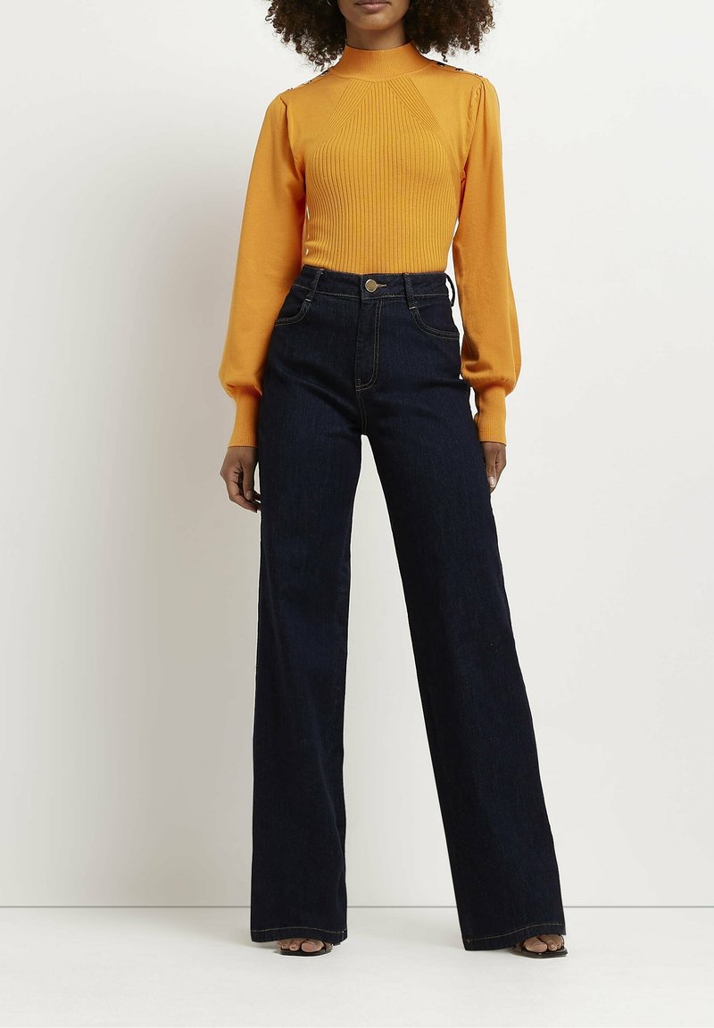 Femme portant un pull côtelé orange vif à manches longues et un jean large taille haute de couleur sombre, debout devant un fond blanc.