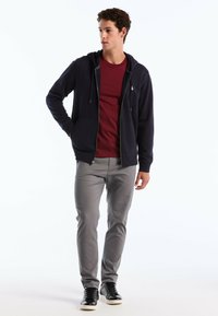 Haut à fermeture éclair marine avec cordons de serrage, pantalon gris ajusté et baskets noires sur un modèle dans un fond neutre. Accentuée par des poignets côtelés.