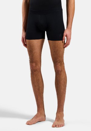 Jambes masculines portant un short noir ajusté, debout pieds nus sur fond blanc, montrant du milieu de la cuisse jusqu'aux pieds et une partie des mains.