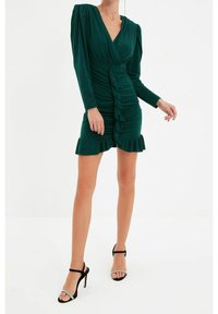 Robe mini verte à manches longues avec un design croisé, devant froncé et ourlet volanté. Fabriquée en tissu lisse, avec une texture et des détails subtils.