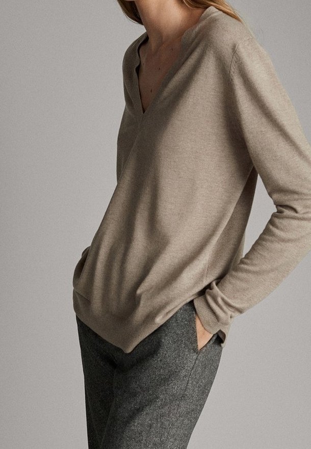 Femme portant un pull beige à col en V et un pantalon en laine gris, les mains dans les poches, debout devant un fond uni.