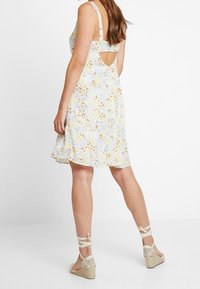 Blommig klänning med ljusblå bas, gula och vita mönster, ärmlös design, öppen ryggdetalj och utsvängd kjol; parat med beige espadriller.