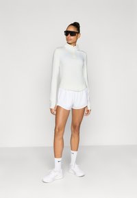 Ελαφρύ, μακρυμάνικο ζακέτο με φερμουάρ σε ανοιχτό πράσινο, σε συνδυασμό με λευκό αθλητικό σορτς. Αξεσουάρ περιλαμβάνουν μαύρα γυαλιά ηλίου και λευκά αθλητικά παπούτσια.