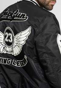 Zwart bomberjack met witte geborduurde patches, waaronder "TopGun," vleugels en het cijfer "23." Satijnachtige textuur met geribbelde kraag.