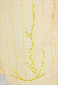 Shorts jaune pâle avec des rayures verticales jaunes. Présente un motif abstrait brodé en fil jaune. Texture lisse et coupe décontractée.