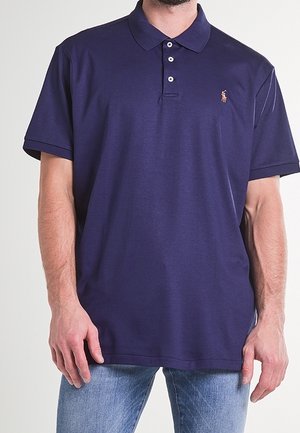 Homme portant un polo bleu foncé à manches courtes avec trois boutons et un jean bleu clair, debout devant un fond uni.