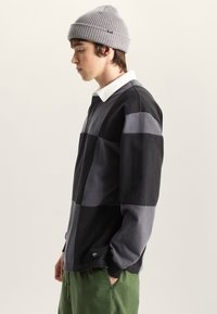 Ung man i grå mössa, svart och grå lapptäcksmönstrad långärmad skjorta med vit krage och gröna byxor, stående i profil med handen i fickan.