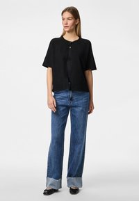 Melnā, īsām piedurknēm džemperī, izgatavots no teksturēta auduma ar apaļu kakla izgriezumu un vienu pogu aizdari, apvienots ar platiem, uzgrieztiem ziliem džinsiem.