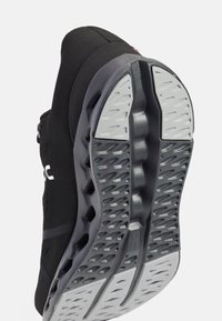 Schwarzer Sportschuh mit einer strukturierten, flexiblen Sohle mit Rillen und einer gepolsterten Zwischensohle. Das Obermaterial besteht aus atmungsaktivem synthetischem Material.
