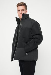 Juodas pūkinis švarkas su aukštu apykakle, užtrauktuku ir stačiakampio formos siuvinėjimu. Yra šoniniai kišeniai ir matinė tekstūra.