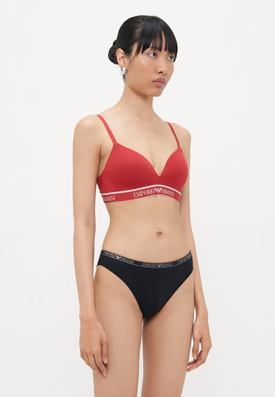 Sarkanais brafiks ar platīgu elastīgu jostu, kurā ir izšuvums "EMPORIO ARMANI" baltā krāsā, komplektā ar melnajiem apakšbiksēm, kurām ir līdzīga josta ar logotipu.