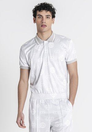 Jonge man met krullend haar die een wit poloshirt met patroon en bijpassende broek met elastische tailleband draagt, staand met één hand in de zak.