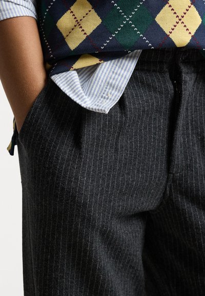 Calças cinzas às riscas com cintura ajustada, combinadas com uma camisa às riscas azul e branca e um colete xadrez multicolorido.