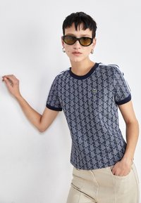 Tílko s námořnickým vzorem a geometrickým designem, s ribovaný límcem a manžetami. Model má na sobě béžové kalhoty a sluneční brýle.