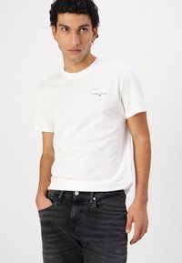 Weißes Baumwoll-T-Shirt mit kurzen Ärmeln, das ein kleines Logo auf der Brust aufweist. Kombiniert mit dunkelgrauen Slim-Fit-Jeans, die Hände in der Tasche.