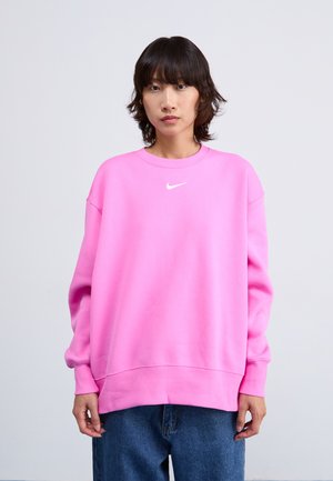 Svetlo roza pulover s krojem na vratu, rebrastimi manšetami in robom. Na prsih ima bel Nike logotip. Usklajen z modrimi kavbojkami.