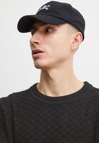 Black cap with embroidered text, worn with a textured dark sweater featuring a checkerboard pattern and a ribbed neckline.