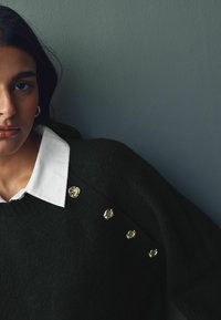 Sort sweater med tekstureret overflade, med en hvid skjorte under og guldhardware detaljer på skuldrene.