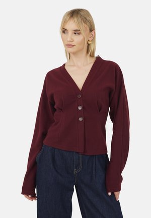 Kobieta w bordowym, zapinanym na guziki kardiganie i ciemnoniebieskich dżinsach, patrząca na bok, z złotymi kolczykami i zrelaksowaną pozą na jednolitym tle.