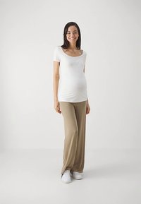 Ένδυμα εγκυμοσύνης που περιλαμβάνει ένα εφαρμοστό λευκό τοπ με λαιμόκοψη στρογγυλή και άνετο μπεζ παντελόνι. Το μοντέλο φορά λευκά αθλητικά παπούτσια, με τα μαλλιά κάτω.