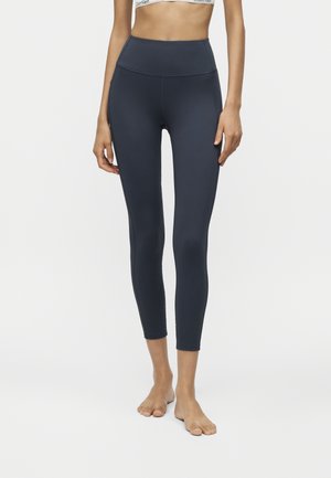 Vrouw draagt zwarte, hoog getailleerde cropped leggings en een witte sportbeha met logoband op een effen lichte achtergrond.