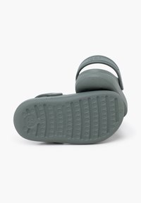 Grüne Gummisandale mit strukturiertem Fußbett, verstellbarem Riemen und einem runden, erhöhten Markierungsdetail. Die Sohle verfügt über ein gemustertes Profil.