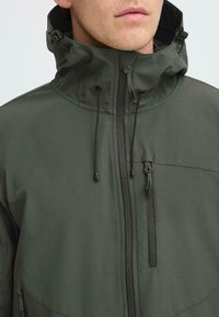 Gros plan sur un homme portant une veste à capuche vert foncé avec un col zippé et une poche sur la poitrine, avec des cordons tombant de la capuche.