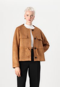 Tan suède jack met een ronde hals, voorzakken en zwarte knopen. Heeft een cropped ontwerp en een strikdetail in de taille.