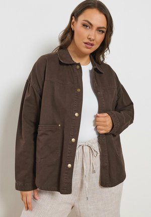 Vrouw die een bruine jas met knopen draagt over een wit shirt en lichte gestreepte broek, poserend tegen een effen witte muur.