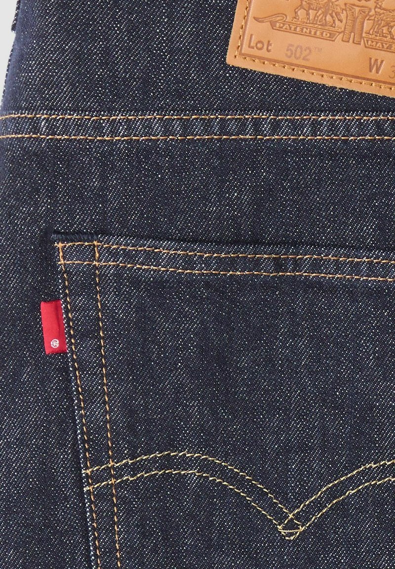 Bolsillo de vaqueros de mezclilla azul oscuro con costuras naranjas, una etiqueta de tela roja y un parche de cuero marrón con texto y gráficos en relieve.