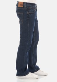 Vaqueros de mezclilla azul oscuro con un corte recto, cinco bolsillos, costuras sutiles y un parche de cuero en la parte trasera de la cinturilla. Zapatillas blancas de deporte usadas.