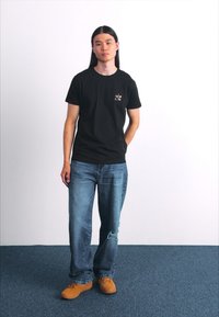 Tricou negru din bumbac cu un grafic colorat mic în partea stângă a pieptului, purtat cu blugi de denim ușor decolorati, de culoare albastră, și pantofi maro.