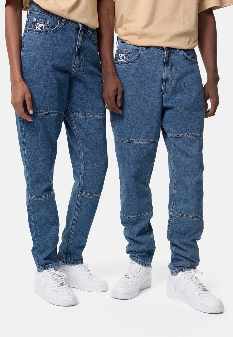 Μπλε τζιν παντελόνι με χαλαρή εφαρμογή, με σχέδιο patchwork, πέντε τσέπες και ελαφρύ φθόρο. Συνδυασμένο με λευκά αθλητικά παπούτσια.