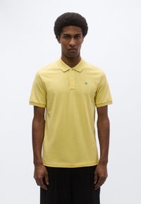 Κίτρινο πόλο πουκάμισο από βαμβάκι, με κλασικό γιακά, κοντά μανίκια και μικρό πράσινο λογότυπο στο στήθος. Απλός, μονόχρωμος σχεδιασμός.