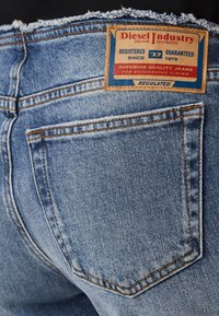 Lys blå denimjeans med en slitt midje, med et brunt lærmerke med blå og rød tekst, og oransje sømdetaljer.