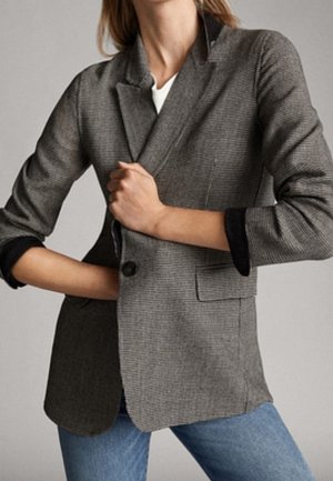 Kvinde iført en grå ternet blazer med sorte opkørte manchetter og blå jeans, der justerer blazeren med den ene hånd.