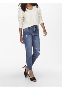 Pull beige clair à col en V, jean bleu déchiré avec des trous, et sandales à talons noirs. Coupe décontractée avec une silhouette ample.