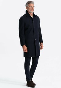 Cappotto di lana blu navy con colletto rialzato, chiusura a cinque bottoni, tasche laterali e silhouette dritta, abbinato a jeans scuri e stivali marroni.