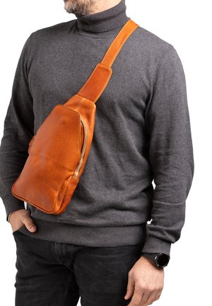 Man met een grijze coltrui en zwarte broek, met een oranje leren crossbodytas en een zwarte smartwatch.