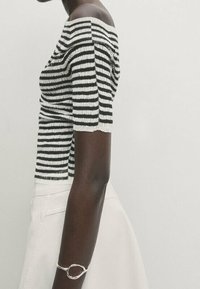 Mörkhynad person som bär en svartvit randig off-shoulder-topp, vit kjol och silverarmband på vänster handled mot en enkel bakgrund.