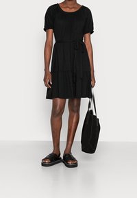 Eine Person steht in einem schwarzen, gestuften Kleid mit kurzen Ärmeln und einem gebundenen Taillengürtel, hält eine schwarze Tasche und trägt schwarze Sandalen.