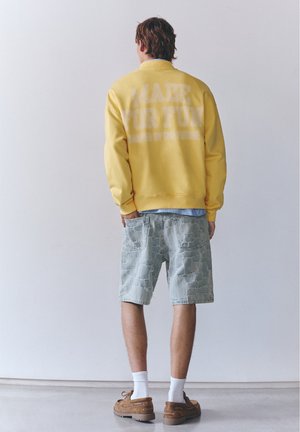 Homme aux cheveux bruns portant un sweat jaune, un short patchwork bleu clair, des chaussettes blanches et des chaussures marron, se tient face à un mur blanc.