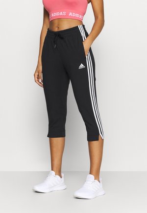 Kobieta ubrana w czarne krótkie spodnie Adidas z białymi paskami, różowy top Adidas i białe trampki na jednolitym tle.